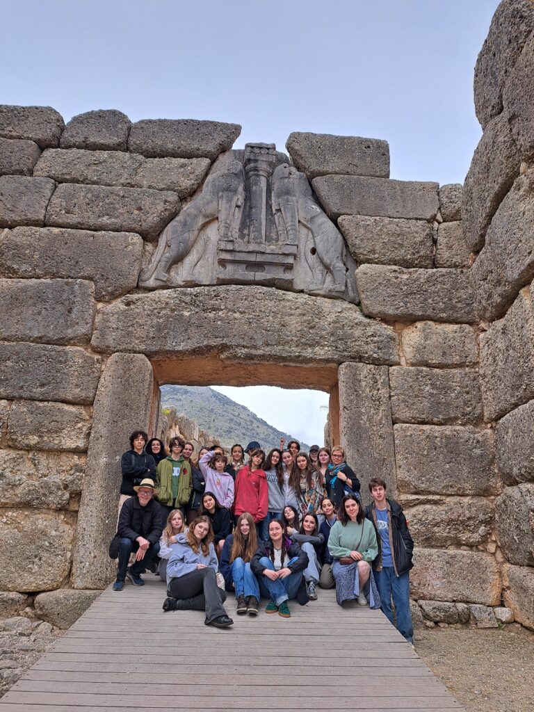 a large group of students from The King Alfred School stand and sit in front of a ruin of an ancient building in Greece