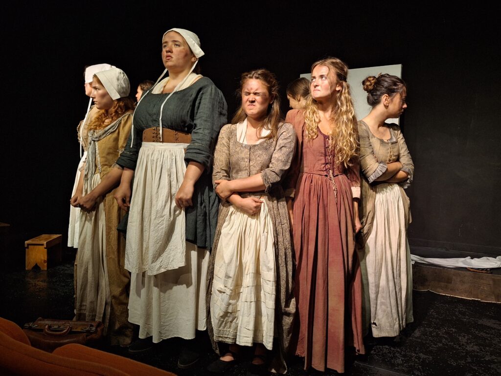 five female students dressed in historical outfits stand on stage during a performance of The Welkin