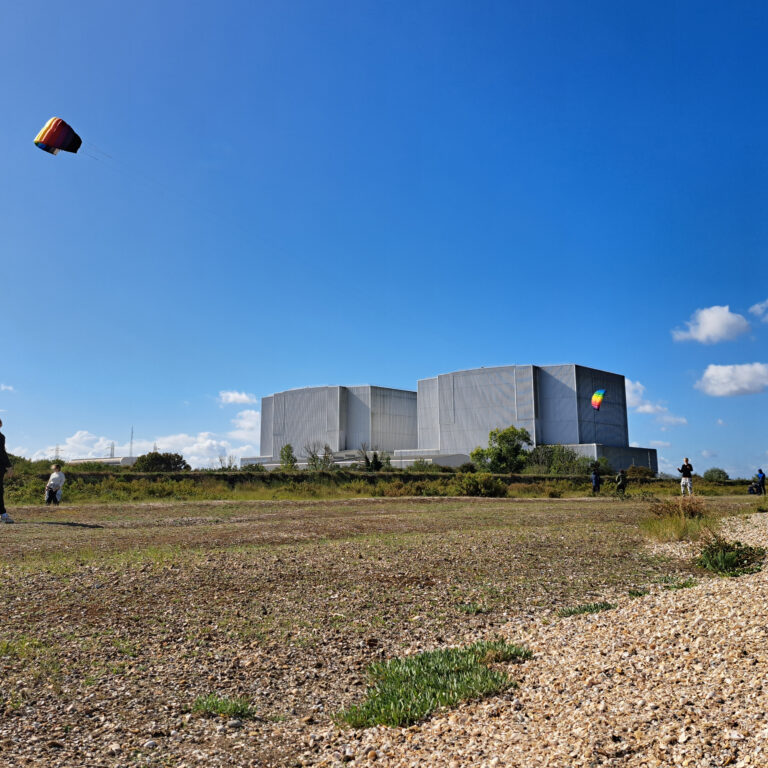 A group of students fly a kite on a vast open space with an industrial building in the background