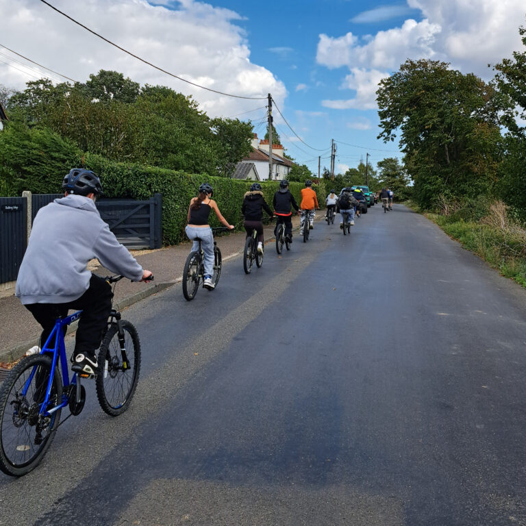 A procession of students cycle along a country road, surrounded by hedges and blue skies.