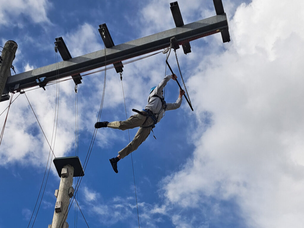 A Sixth Form student leaps through the air into a trapeze bar as part of a high wire course, set against a blue sky with white clouds.