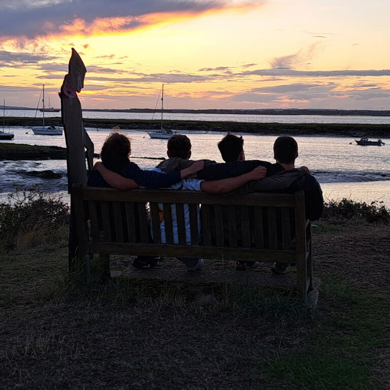 Four boys sit on a bench facing the sea. The sun is setting and they are in shadow.