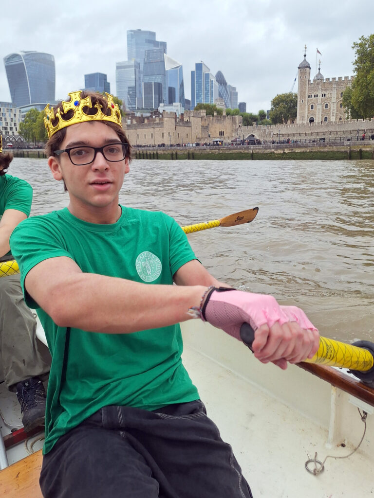 A male student holds an oar inside a wooden row boat on the River Thames. The Tower of London can be seen behind him.