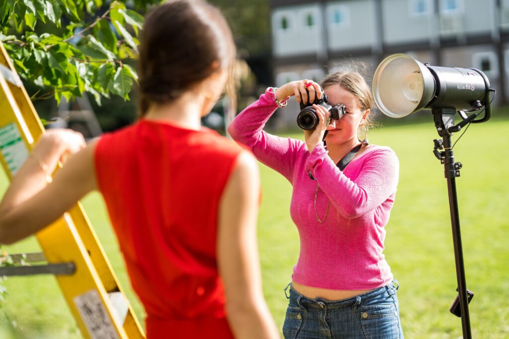 a student in a pink jumper and blue jeans holds a camera and stands beside a light while taking a photo of another student seen from behind wearing a red top