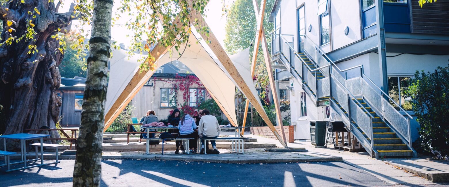 A group of students at The King Alfred School sit and study under a wooden and canvas structure in the school grounds
