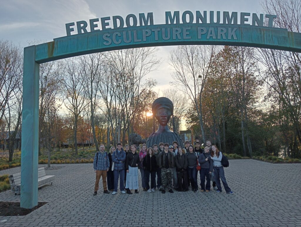 a large group of students along with two teachers stand in front of the Freedom Monument in the USA