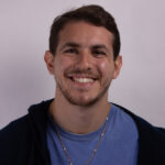 Headshot of a man smiling and wearing a blue tshirt and dark hoodie