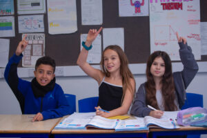 Three students are sat behind a desk in a classroom, one boy and two girls, two of them have their hands raised