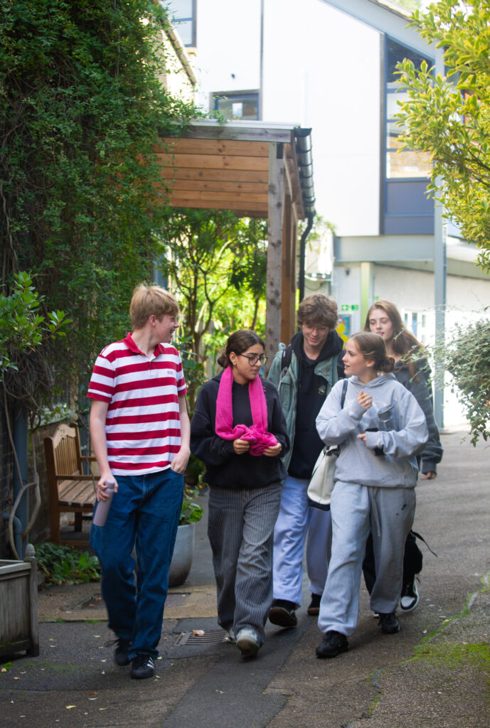 a mixed group of Sixth Form students dressed in casual clothes walk past buildings between lessons at The King Alfred School