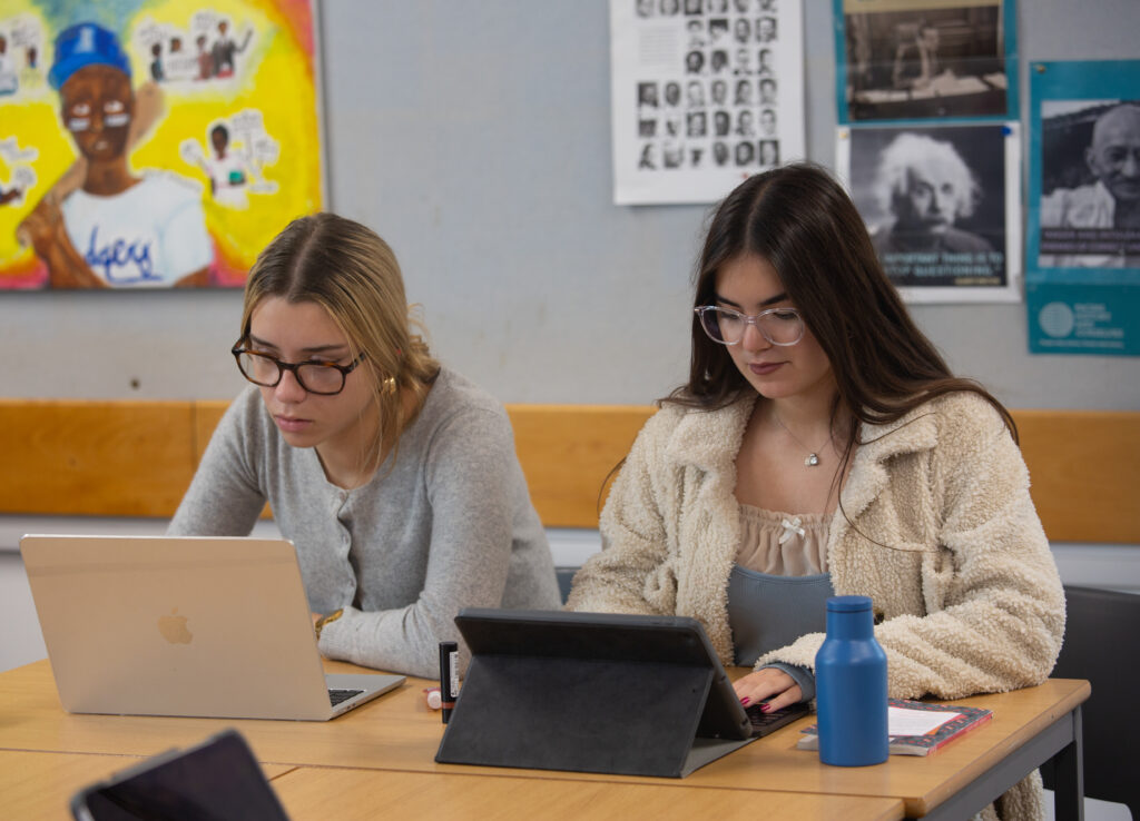 two female students sit at a desk working on laptop computers