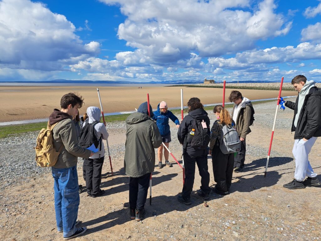 a group of students stand on a british beach holding red and white striped poles and listening to a teacher