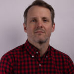Headshot of a man with short dark hair wearing a black and red checked shirt