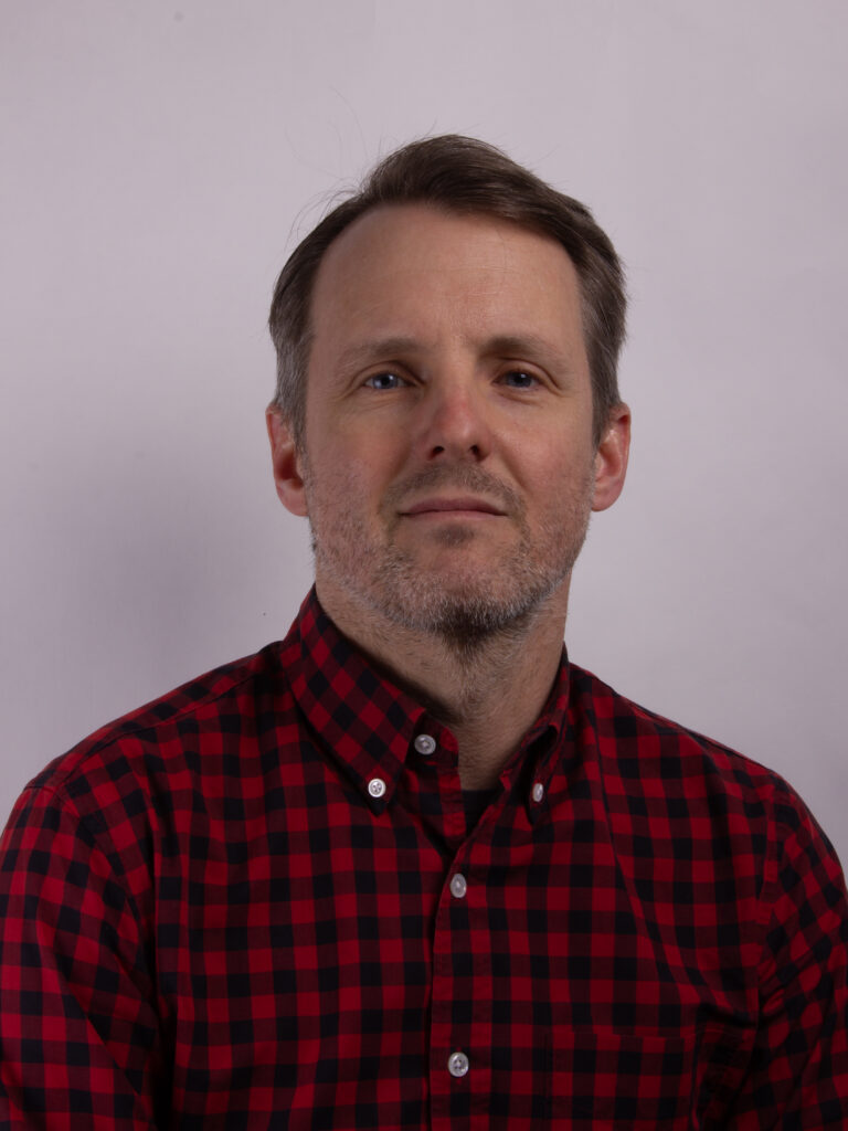Headshot of a man with short dark hair wearing a black and red checked shirt