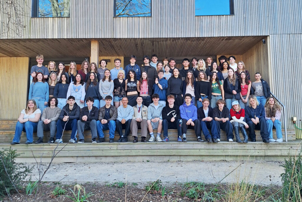 Around 55 Sixth Form students from The King Alfred School stand in three rows posing for a year group photograph outside of the wood-panelled Sixth Form building.