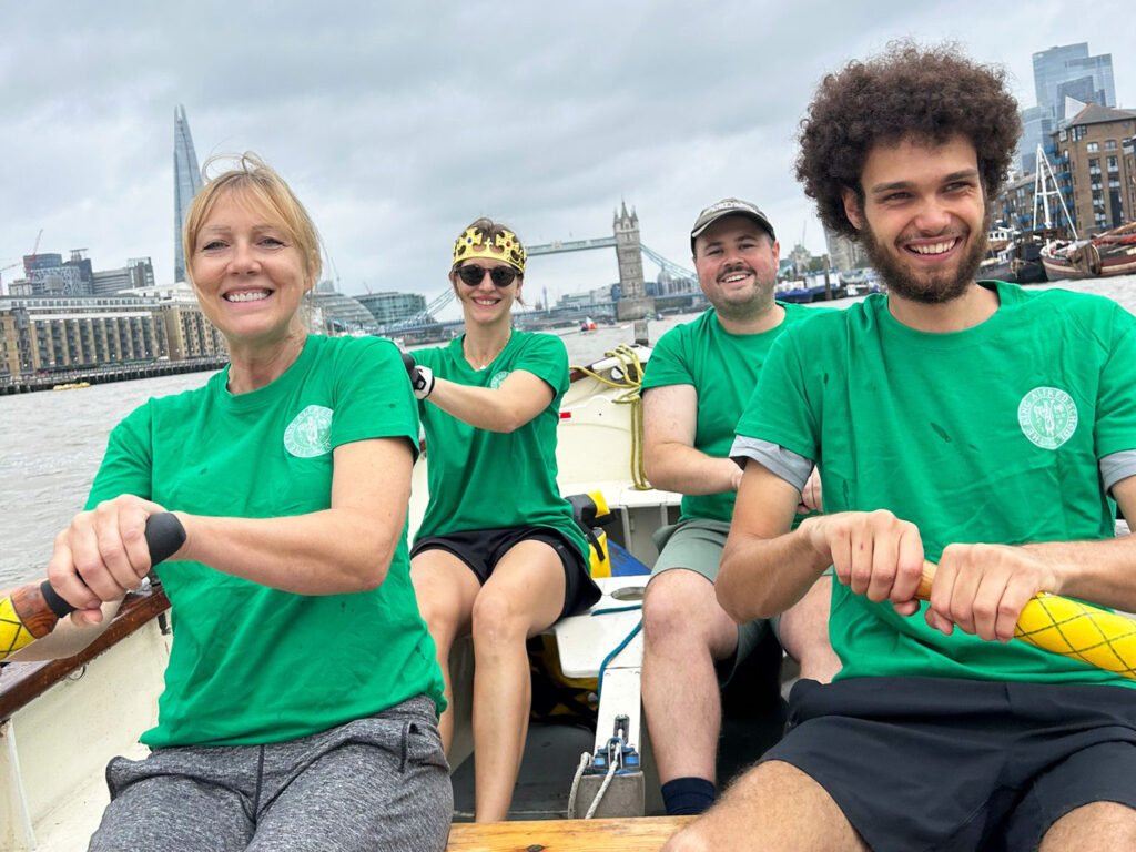 A crew of two women and two men are pictured smiling inside a wooden row boat on the River Thames. Tower Bridge and The Shard are visible in the distance behind them.