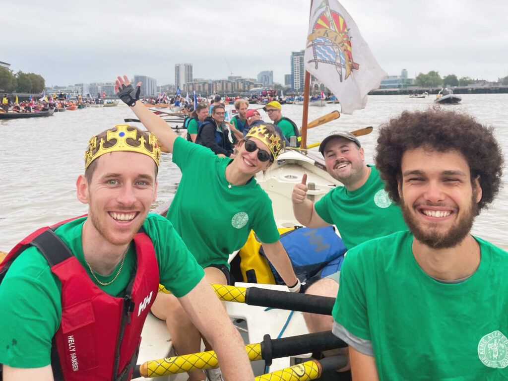 A crew of three men and one woman in a wooden row boat on the River Thames. They are all wearing green t-shirts bearing the King Alfred School logo. There is another crew behind them.