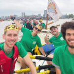 A crew of three men and one woman in a wooden row boat on the River Thames. They are all wearing green t-shirts bearing the King Alfred School logo. There is another crew behind them.