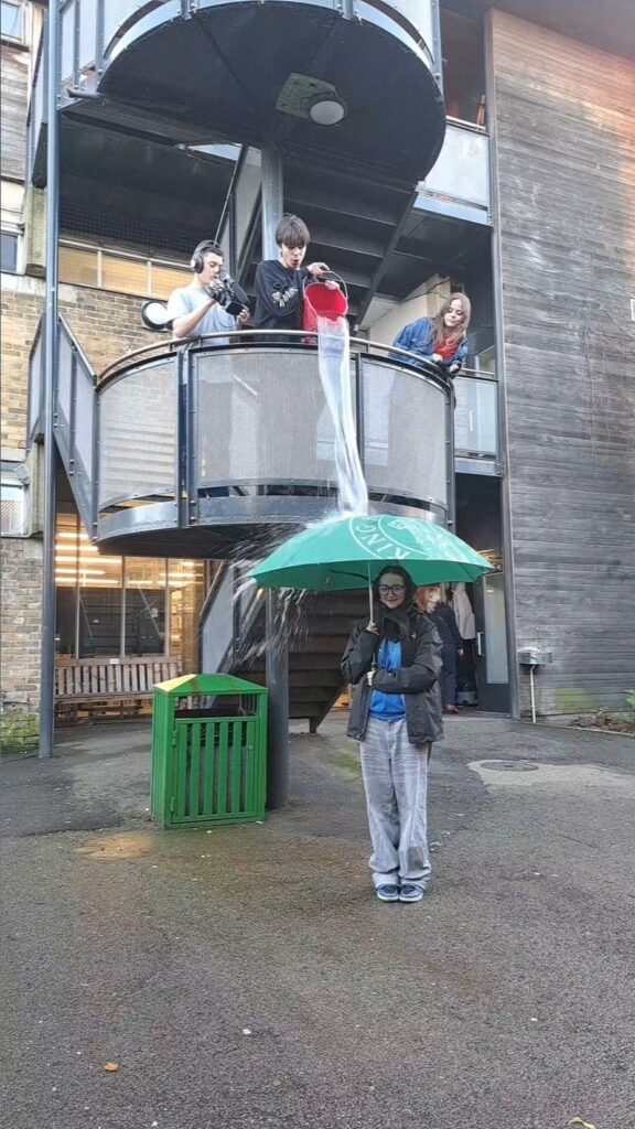 three students stand on a balcony, one is filming while another pours a bucket of water over a student stood below holding a green umbrella