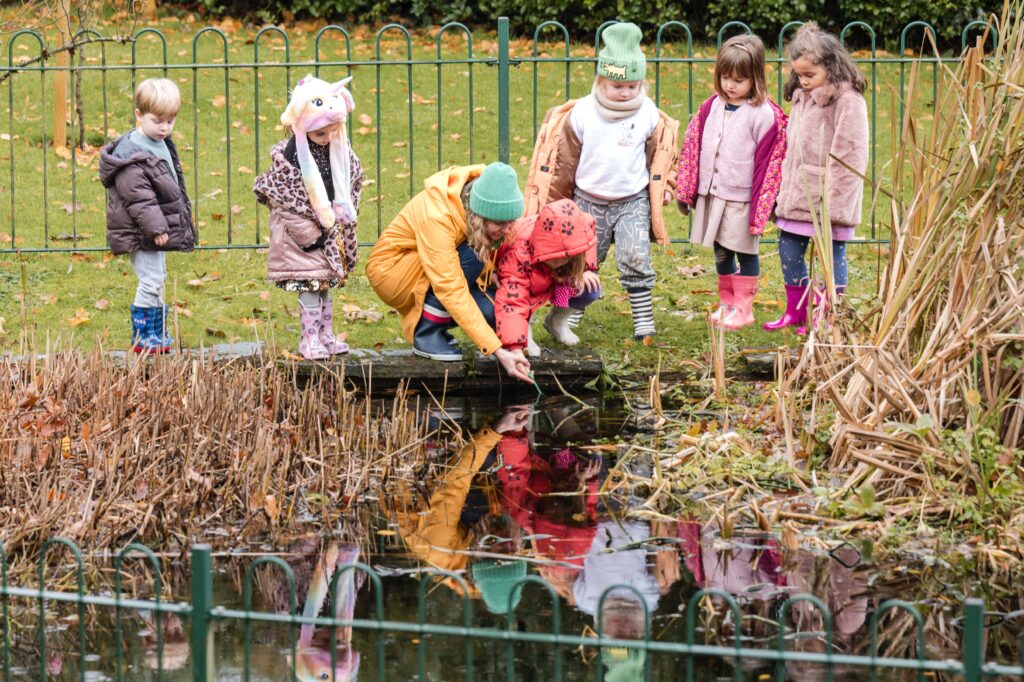 A group of children wearing winter coats examine wildlife in a pond with a teacher