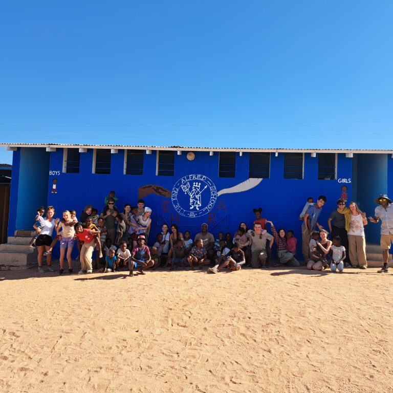 Year 11 students from The King Alfred School gathered in front of a bright blue school building with painted artwork and text on the wall. The building has separate entrances labeled “Boys,” “Girls,” and “Teacher.” The ground is sandy, and the sky is clear and sunny.