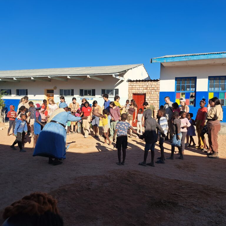 A group of people and children gathered in a sandy courtyard between two school buildings painted white and blue. One building has colorful square decorations on its windows, and the other has a red door and a small brick section. The group appears to be engaged in an outdoor activity under a bright blue sky.