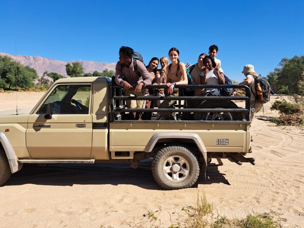 A group of Year 11s standing and sitting in the open back of a beige pickup truck parked on sandy terrain. They are wearing casual outdoor clothing and carrying backpacks. The background shows desert vegetation and distant mountains under a clear blue sky.