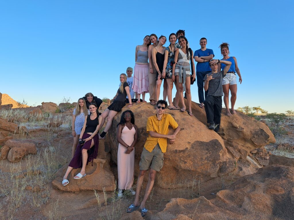 A group of Year 11 students gathered on and around a large reddish-brown rock formation in a desert landscape. The sky is bright blue, and the terrain is dry with scattered vegetation.