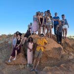 A group of Year 11 students gathered on and around a large reddish-brown rock formation in a desert landscape. The sky is bright blue, and the terrain is dry with scattered vegetation.