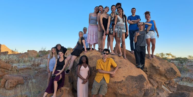 A group of Year 11 students gathered on and around a large reddish-brown rock formation in a desert landscape. The sky is bright blue, and the terrain is dry with scattered vegetation.
