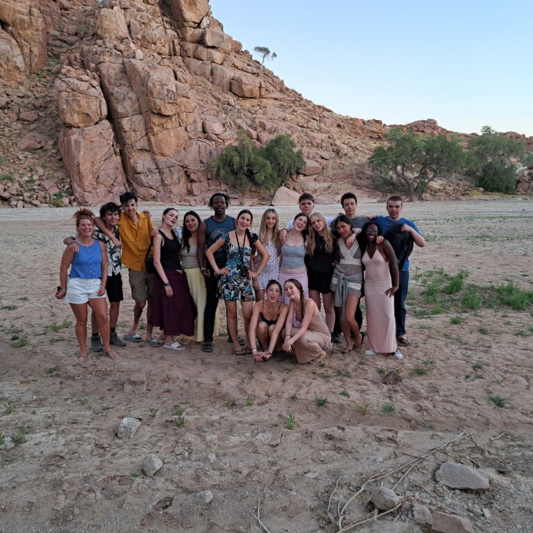 A large group of Year 11 students from The King Alfred School standing together on sandy ground in front of towering rocky cliffs and sparse greenery. The scene is set in a desert environment with clear blue skies overhead.