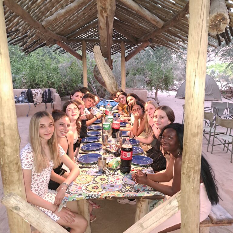 A group of people seated around a long, colorful table under a rustic wooden shelter in an outdoor setting. The table is set with blue plates and drinks, including bottles of Coca-Cola and Sprite. Surrounding the shelter are trees and sandy ground, with camping gear visible in the background.