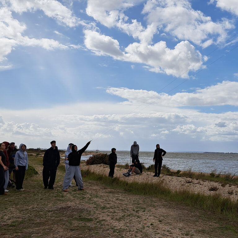 A group of students fly kites along a coastline, with a blue sky behind them.