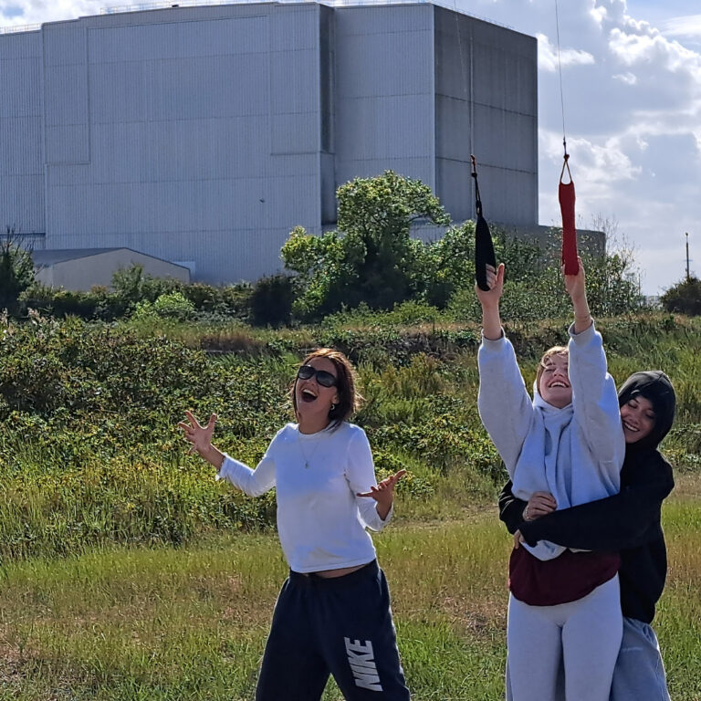Three female Sixth Form students have fun on some grass outside of an industrial building. Two are holding kite strings, and the other is lifting one of her friends up.