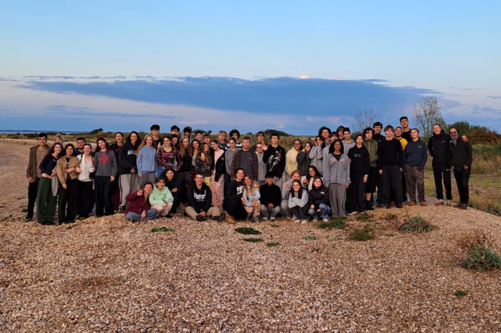 A group photo of approximately 65 Year 12 students from The King Alfred School on a pebble beach at dusk.