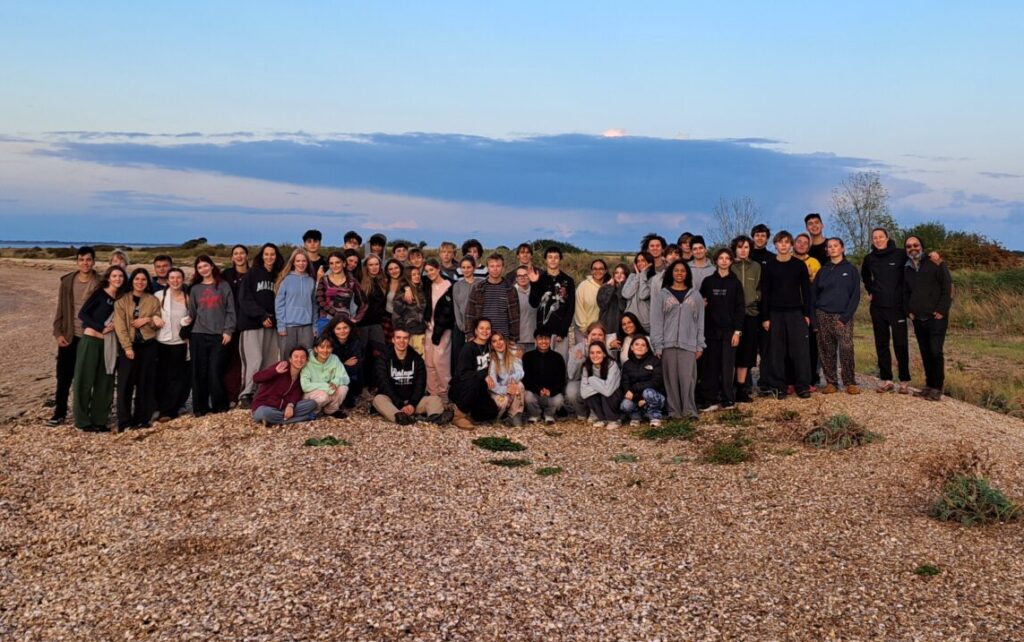 A group photo of approximately 65 Year 12 students from The King Alfred School on a pebble beach at dusk.