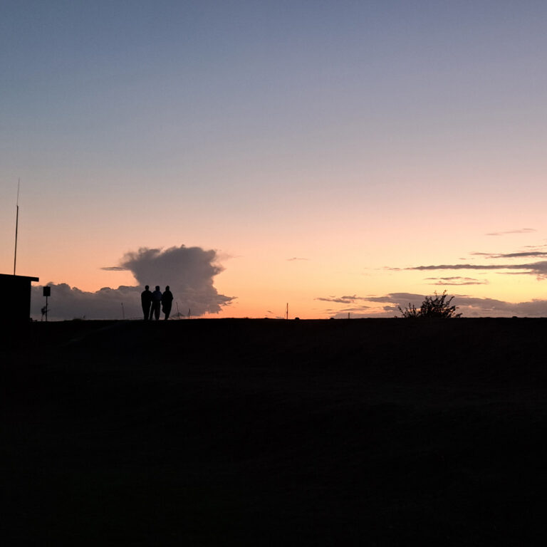 A horizon in shadow at sunset shows the silhouette of small groups of students and a building. The sky is orange, yellow and pale blue.