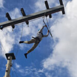 A Sixth Form student leaps through the air into a trapeze bar as part of a high wire course, set against a blue sky with white clouds.
