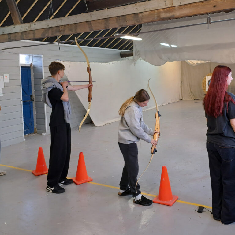 Four students stand in a line at an archery centre, holding crossbows and looking towards a row of targets.