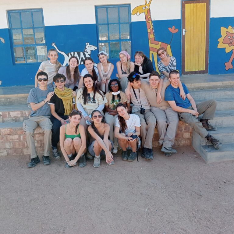 Year 11 students from The King Alfred School posing in front of a bright blue school building with painted artwork and text on the wall. The building has separate entrances labeled “Boys,” “Girls,” and “Teacher.” The ground is sandy, and the sky is clear and sunny.