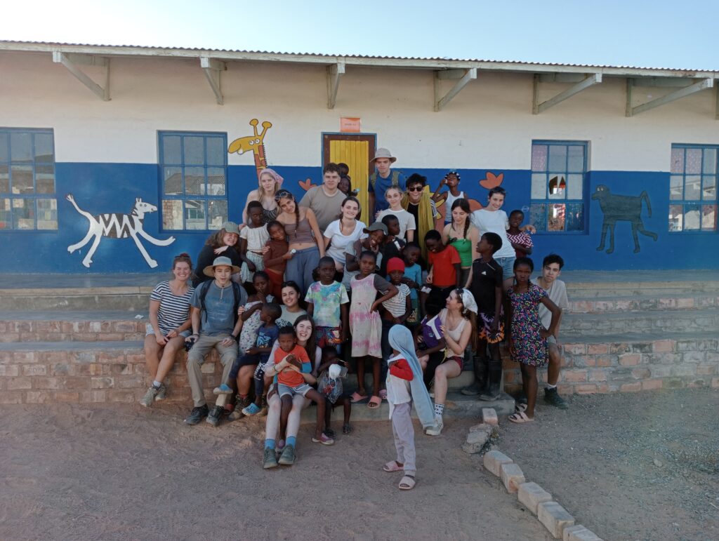 Students from The King Alfred School posing with local children on the steps of a school building. The building has a white and blue painted wall featuring animal illustrations, including a zebra, giraffe, and dog. The group is sitting and standing together in front of two blue-framed windows and a yellow door.