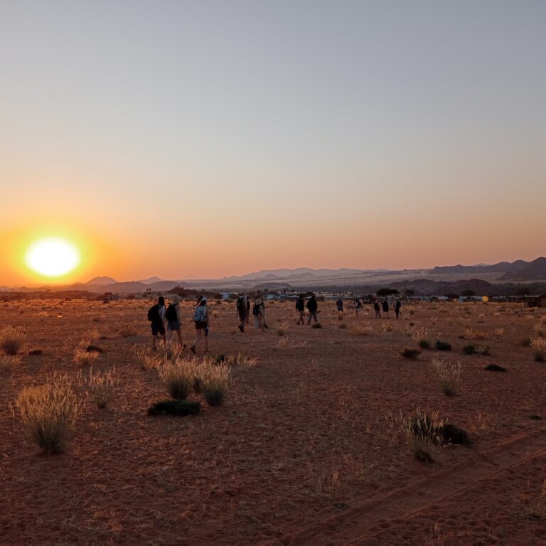 A group of Year 11 students walking across a wide, open desert landscape during sunset. The sun is low on the horizon, casting a warm orange glow over the sandy terrain dotted with sparse vegetation. Mountain ranges are visible in the distance under a clear sky.