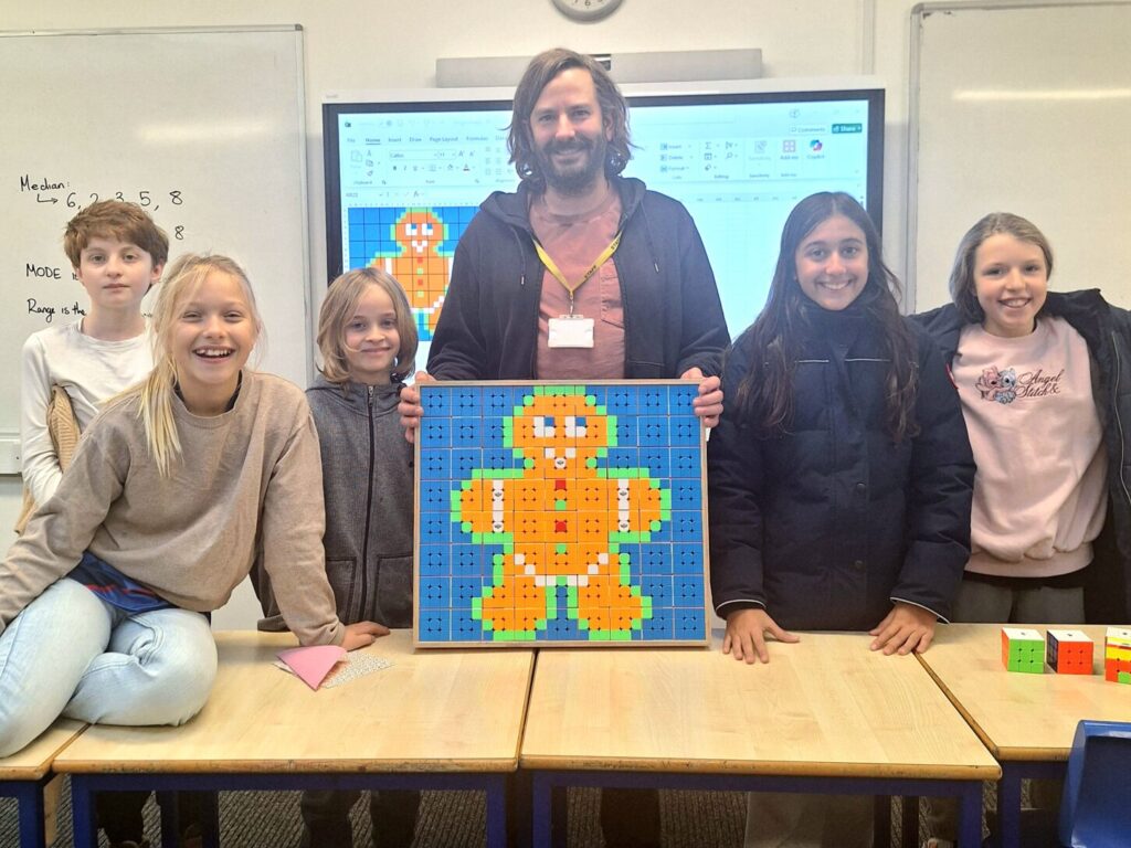 A male student stands behind a desk, flanked by two male and three female students, and holding a gingerbread picture made entirely out of Rubik's cubes