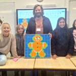 A male student stands behind a desk, flanked by two male and three female students, and holding a gingerbread picture made entirely out of Rubik's cubes