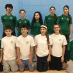 A group of students pose together in a sports hall, arranged in two rows. The back row wears green sports tops, and the front row wears white tops, both bearing The King Alfred School's logo. They are standing and kneeling on a wooden sports floor with a blue wall behind them.
