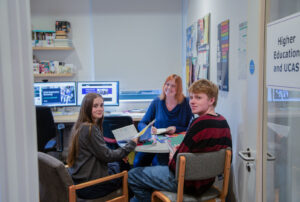 Two students sit at a desk in a teacher's office looking over their shoulders. The teacher is sat on the other side of the desk, looking forwards.