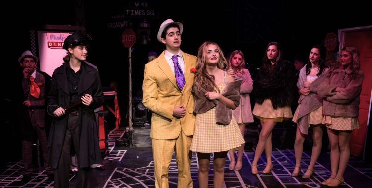 A stage scene from a theatrical production featuring several performers in costume. At the centre, one performer wears a bright yellow checked suit with a purple tie and white hat, standing arm-in-arm with another performer in a yellow gingham dress and a brown fur stole. Other performers in similar yellow dresses and fur stoles stand in the background. The stage floor has a street-map design, and the backdrop includes street signs for Times Square, Broadway, and a stop sign, as well as a diner sign reading “Mindy’s Diner.”