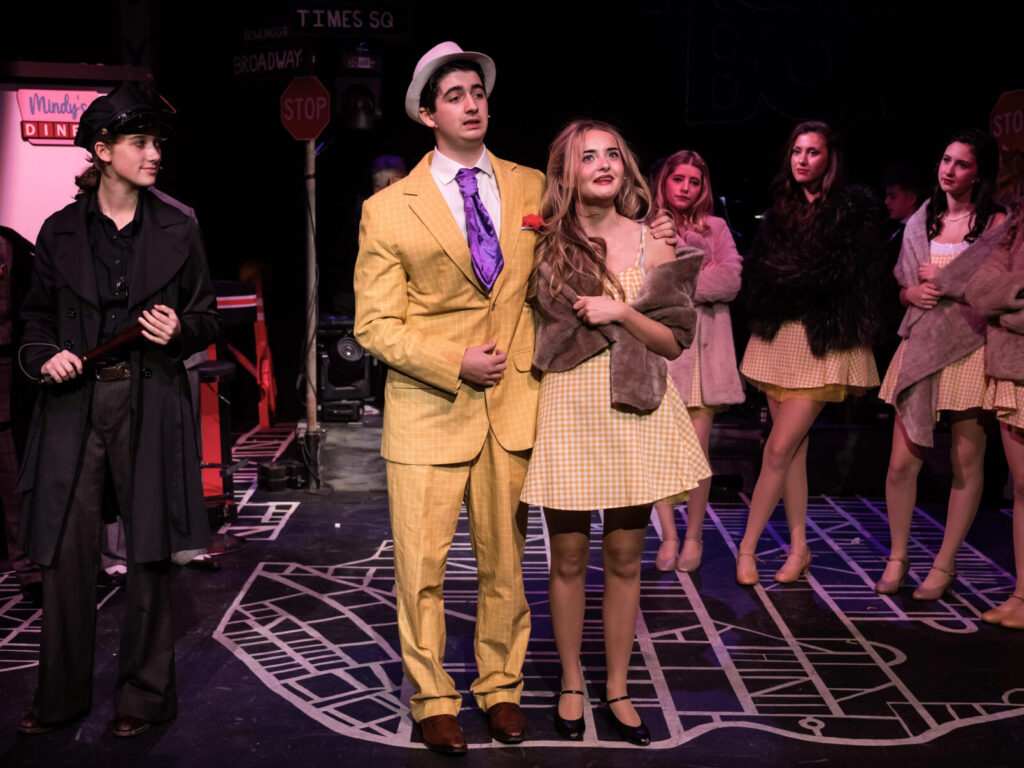 A stage scene from a theatrical production featuring several performers in costume. At the centre, one performer wears a bright yellow checked suit with a purple tie and white hat, standing arm-in-arm with another performer in a yellow gingham dress and a brown fur stole. Other performers in similar yellow dresses and fur stoles stand in the background. The stage floor has a street-map design, and the backdrop includes street signs for Times Square, Broadway, and a stop sign, as well as a diner sign reading “Mindy’s Diner.”