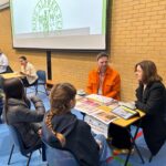 A row of desks in a sports hall, with students sat on one side and parents on the other. They are engaged in conversation, with labels on each desk detailing the industry the parent works in, including architecture.