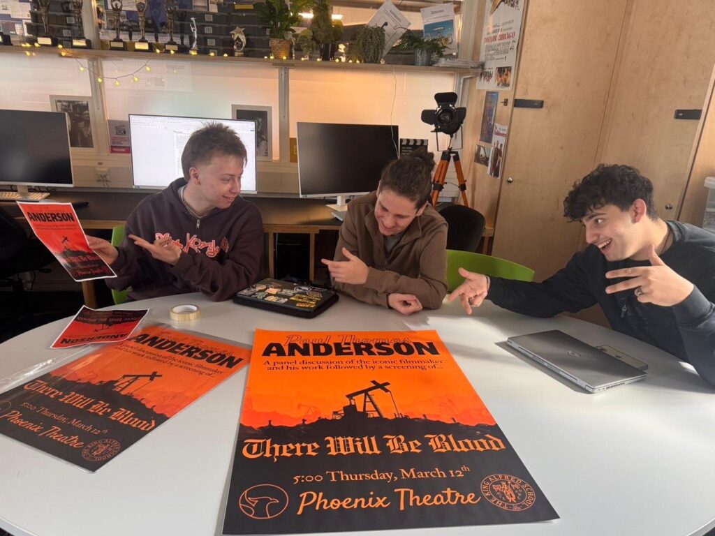 Three male film students sit at a a classroom table, holding and pointing at posters for an event they have organised. The poster reads: 'Paul Thomas Anderson. A panel discussion of the iconic filmmaker and his work followed by a screening of There Will Be Blood. Thursday 12 March at The King Alfred Phoenix theatre'. It features a blood orange background with a hill and oil derrick silhouetted in black.