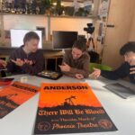 Three male film students sit at a a classroom table, holding and pointing at posters for an event they have organised. The poster reads: 'Paul Thomas Anderson. A panel discussion of the iconic filmmaker and his work followed by a screening of There Will Be Blood. Thursday 12 March at The King Alfred Phoenix theatre'. It features a blood orange background with a hill and oil derrick silhouetted in black.